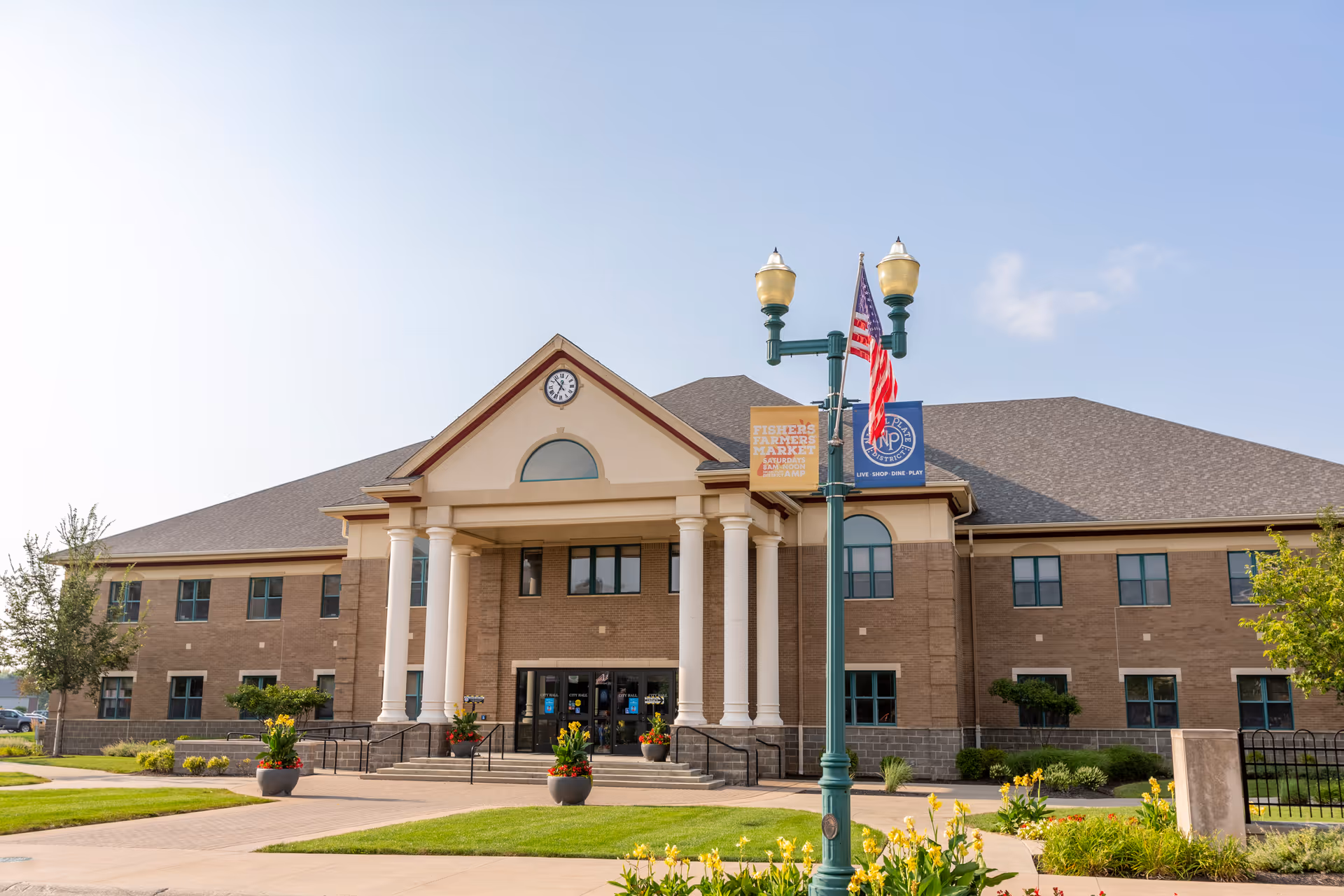 Front exterior view of a large two-story brick building with a peaked roof and white columns at the entrance. There is a clock above the entrance and a lamp post with an American flag and banners in front of the building. The surrounding area has green grass, flowers, and a clear sky.