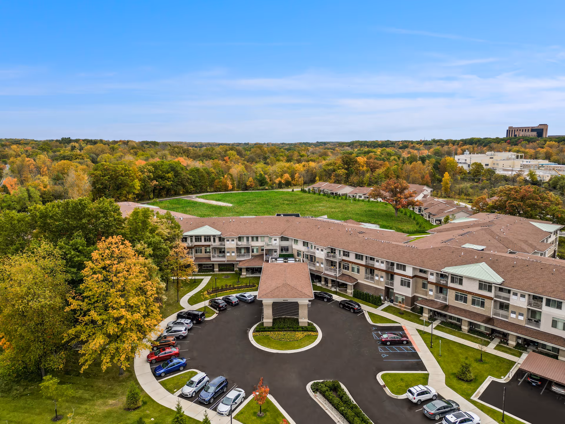 Aerial view of All Seasons Ann Arbor senior living facility showing a large, U-shaped building surrounded by trees with autumn foliage. The building has a covered entrance with a circular driveway and several parked cars. The background features a forested area and a clear blue sky.