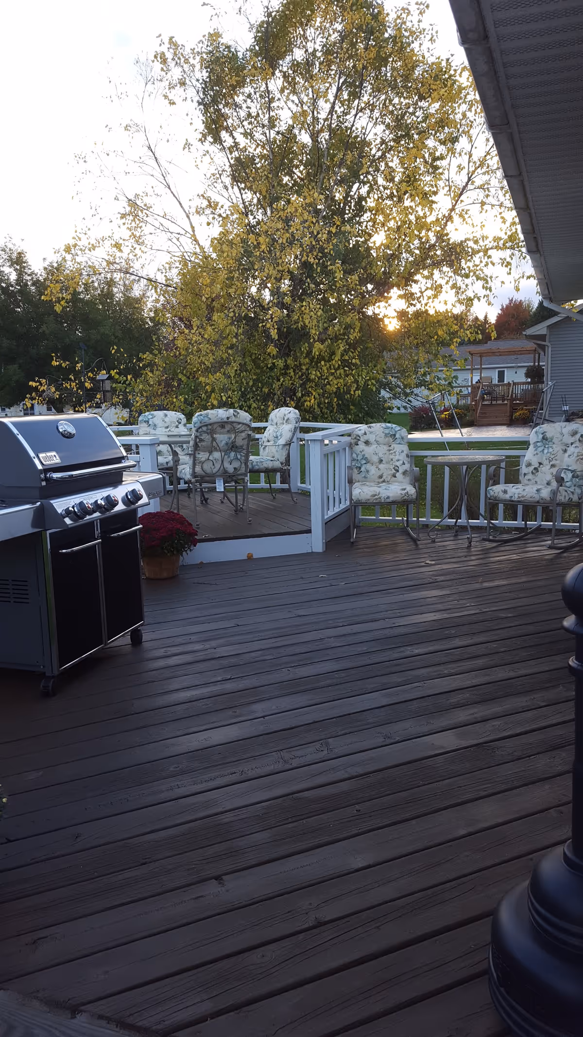 Outdoor wooden deck area with a barbecue grill on the left side, several cushioned chairs with floral patterns around small tables, a potted plant with red flowers, and a large tree with yellowing leaves in the background. Residential houses and a lawn are visible beyond the deck.