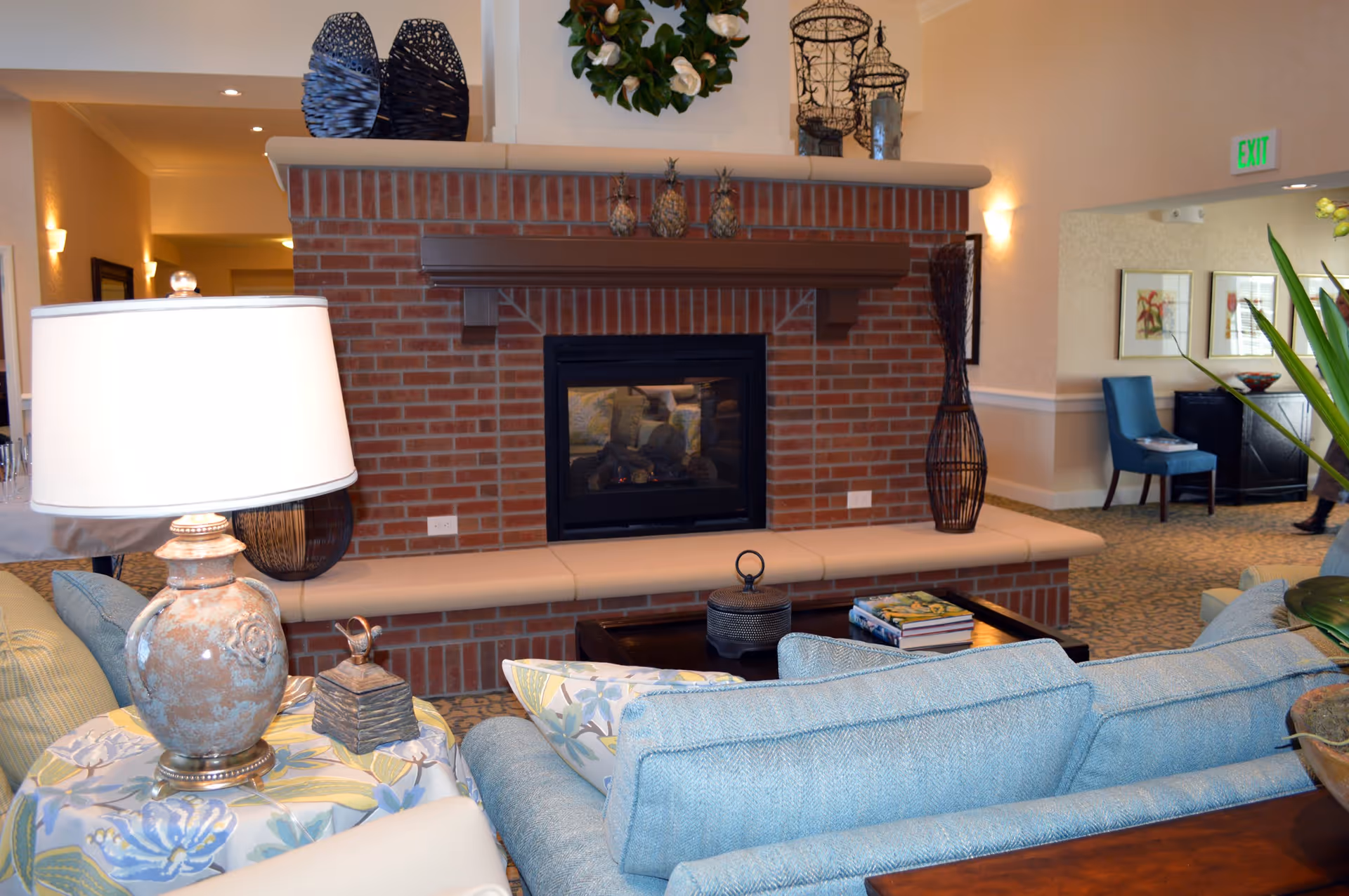 A cozy senior living facility common area featuring a brick fireplace with decorative pineapples on the mantel. In front of the fireplace is a light blue sofa with floral patterned pillows, a side table with a lamp, and a coffee table with books and a decorative item. The room has warm lighting and tasteful decor including a wreath above the fireplace and framed artwork on the walls.