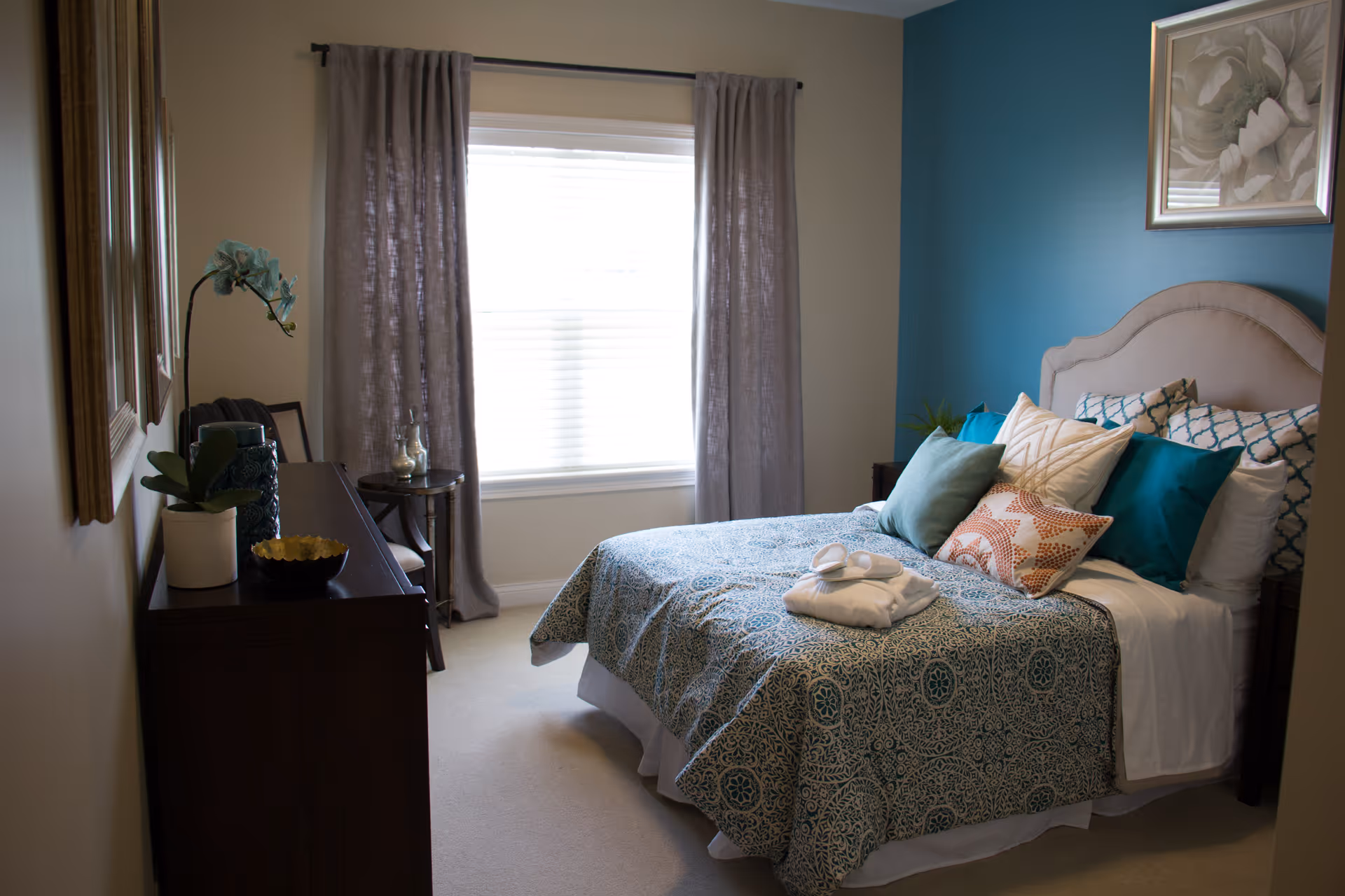 A cozy bedroom with a bed covered in a patterned blue and white bedspread, multiple decorative pillows, and a folded white robe placed on top. The room has a blue accent wall behind the bed, a window with gray curtains letting in natural light, a wooden dresser with a potted plant and decorative items, and framed artwork on the walls.