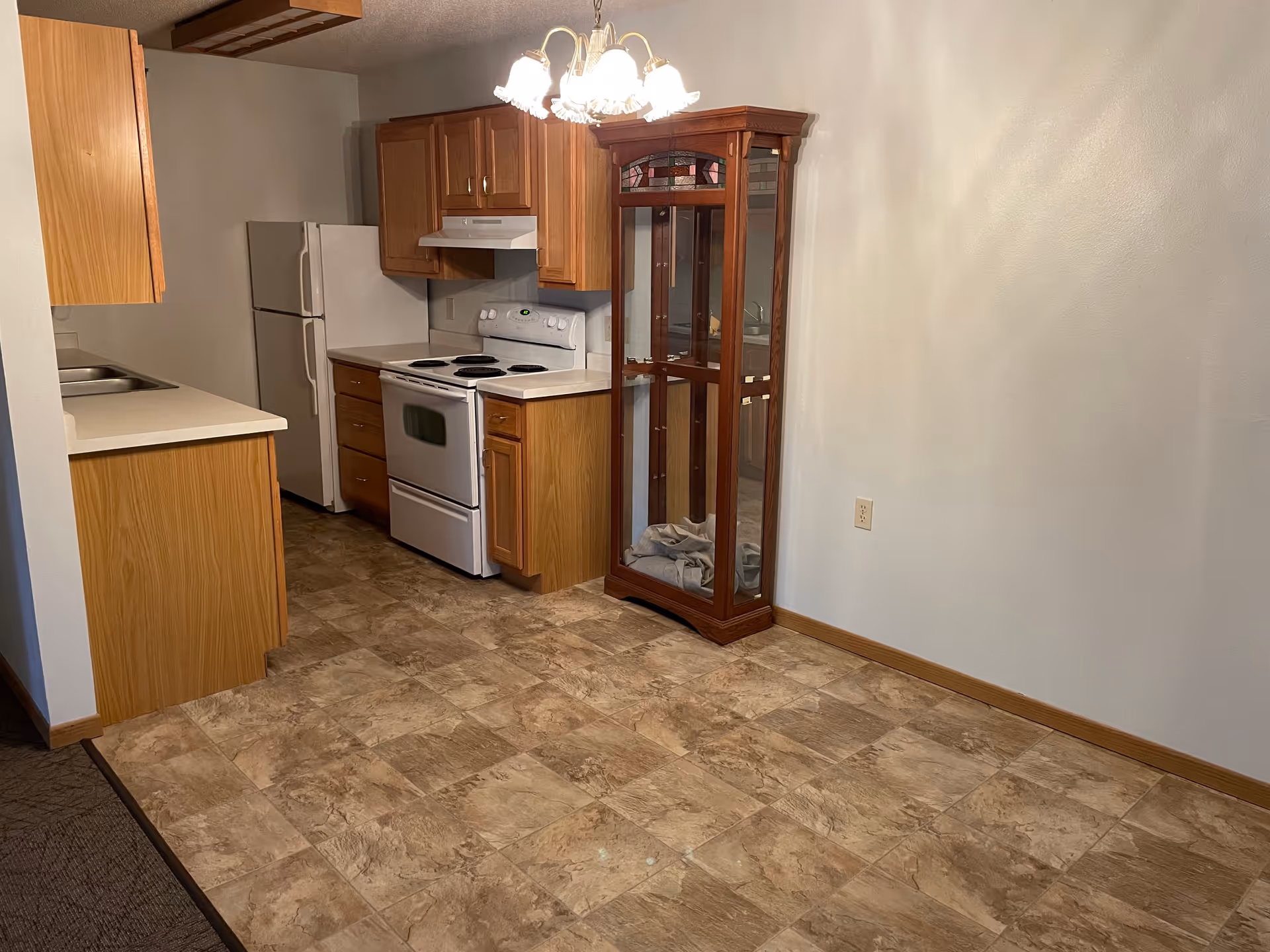 Interior view of a kitchen area with wooden cabinets, a white refrigerator, a white stove with an overhead vent, and a glass display cabinet with wooden frame. The floor is covered with beige and brown patterned tiles, and there is a chandelier light fixture hanging from the ceiling. The walls are painted light beige.