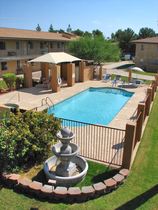 Outdoor swimming pool area at a senior living facility with a small fountain in the foreground, surrounded by a low brick border and greenery. The pool is fenced with a gate, and there are lounge chairs and a shaded seating area nearby. Apartment buildings are visible in the background under a clear blue sky.