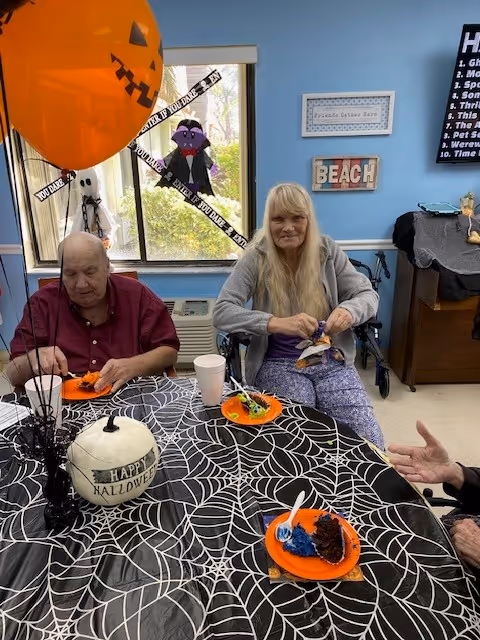 Two elderly residents sit at a Halloween-decorated table eating dessert in a common room.