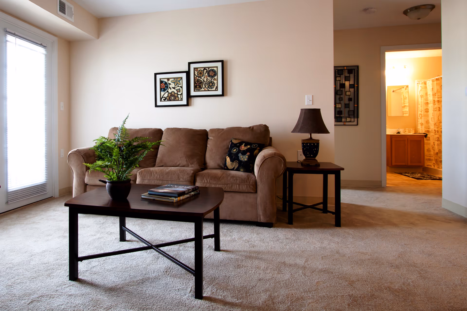 A cozy living room with a brown sofa adorned with a decorative pillow, a dark wooden coffee table with a potted plant and books, and a side table with a lamp. Two framed floral artworks hang on the wall above the sofa. To the right, an open doorway leads to a brightly lit bathroom with a shower curtain and vanity visible. Natural light enters through a window with blinds on the left side.