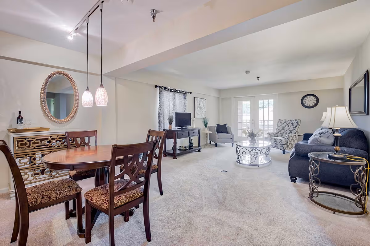 A bright and spacious living and dining area in a senior living facility. The dining area features a wooden table with four upholstered chairs, two pendant lights hanging above, and a decorative mirror on the wall. The living area includes a dark sofa, two patterned armchairs, a round glass coffee table, a side table with a lamp, a TV on a stand, and a wall clock. Large windows and glass doors allow natural light to fill the room.