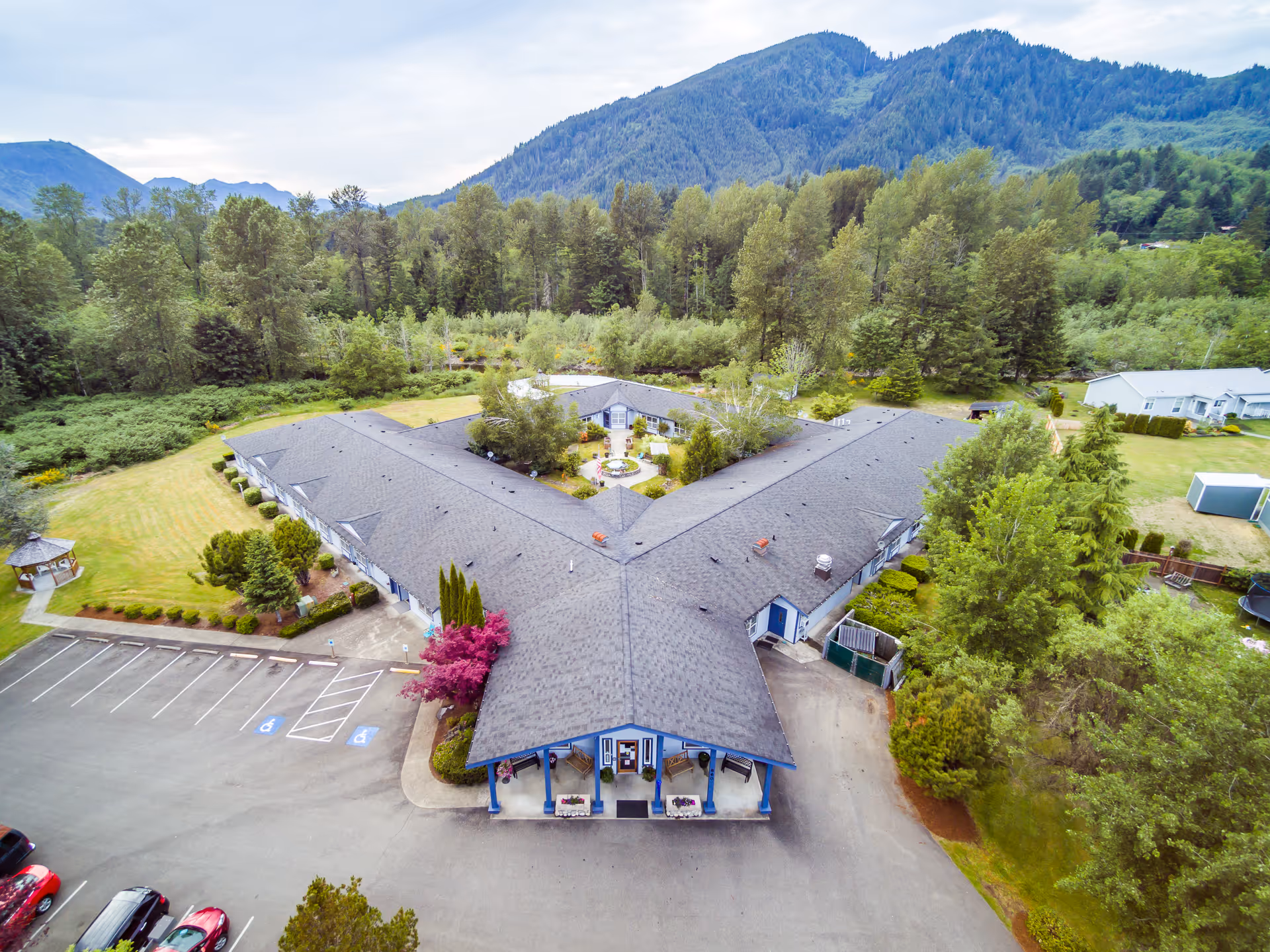 Aerial view of Heritage House Morton Assisted Living and Memory Care facility showing a Y-shaped building surrounded by trees, greenery, and mountains in the background. The building has a gray roof and blue accents, with a parking lot visible in the foreground.