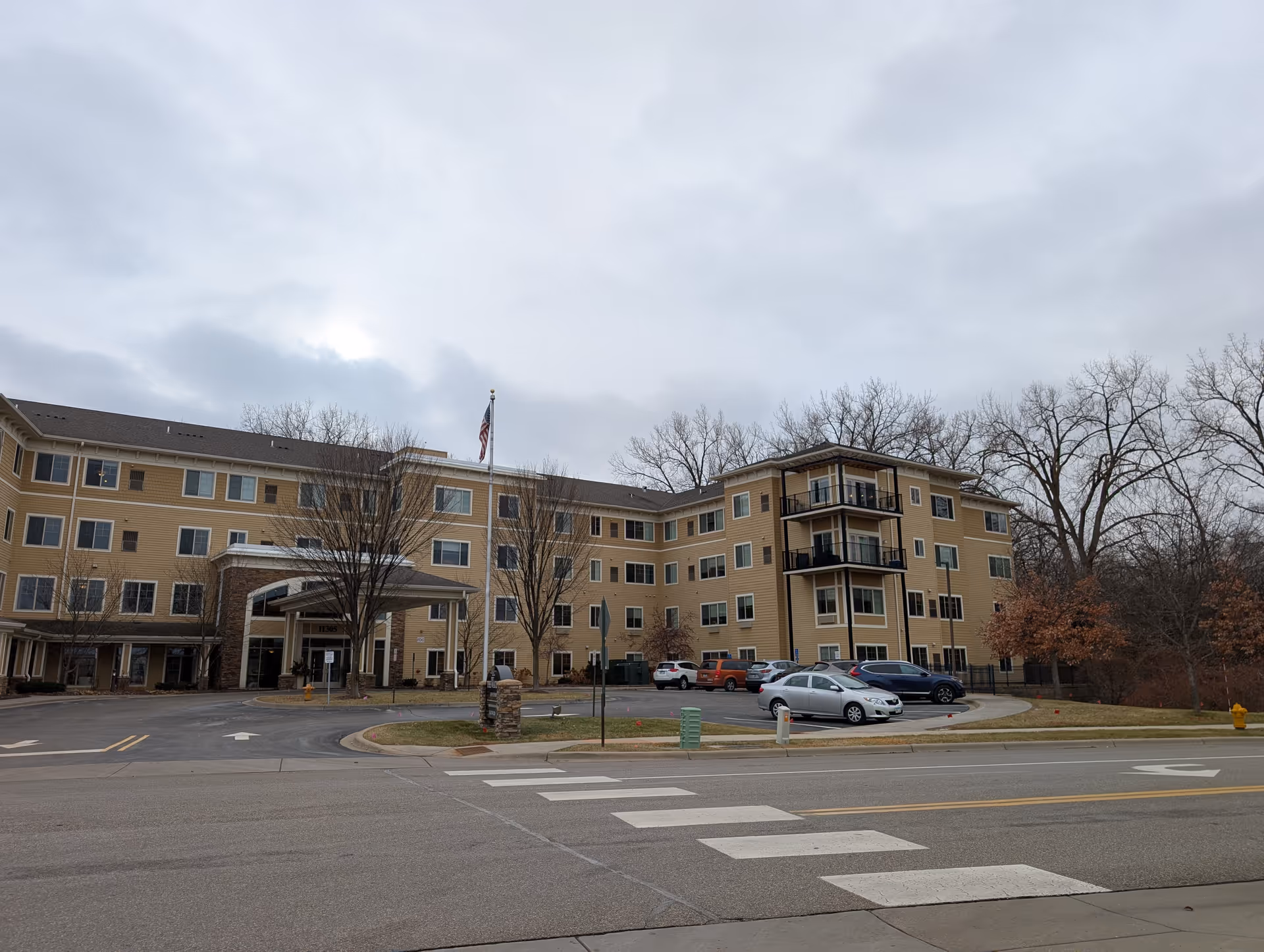 Four-story beige senior living building with a covered entrance, parked cars, an American flag, and leafless trees under a cloudy sky.