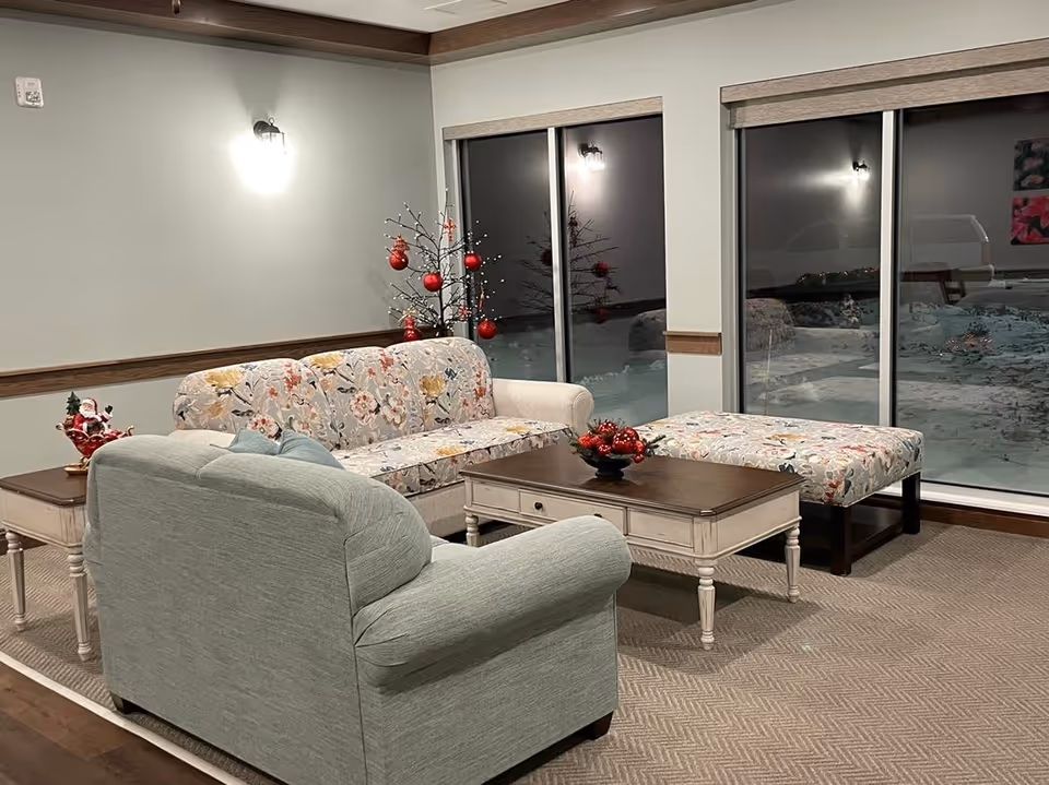 A cozy living room area with a floral-patterned sofa, a light green armchair, a matching floral ottoman, and a wooden coffee table decorated with red Christmas ornaments. There is a small decorative tree with red baubles in the corner near large windows showing a snowy outdoor scene at night.
