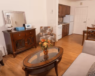 A cozy living room area with a wooden coffee table holding a vase of flowers, a patterned armchair, a wooden cabinet with a mirror and decorative items, and a small kitchen area with wooden cabinets, a stove, and a refrigerator in the background.