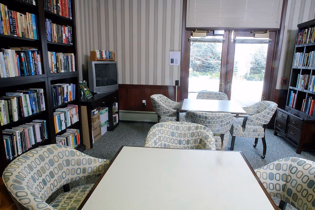 A cozy reading or activity room with patterned armchairs around tables, bookshelves filled with books, a television on a stand, and large glass doors letting in natural light with a view of trees outside.