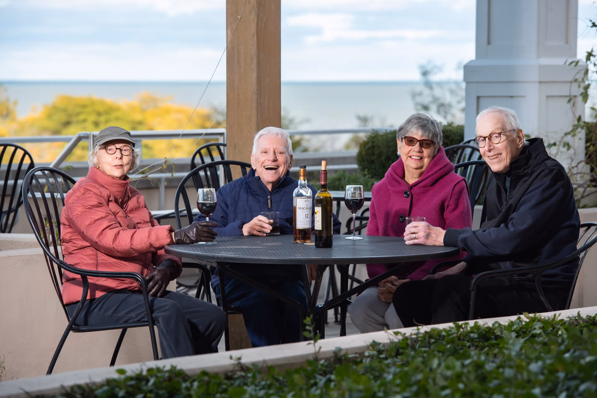 Four elderly people sitting around a round outdoor table on a patio, enjoying drinks with two bottles of wine on the table. They are dressed warmly with jackets and gloves, and there is a scenic view of trees and water in the background.