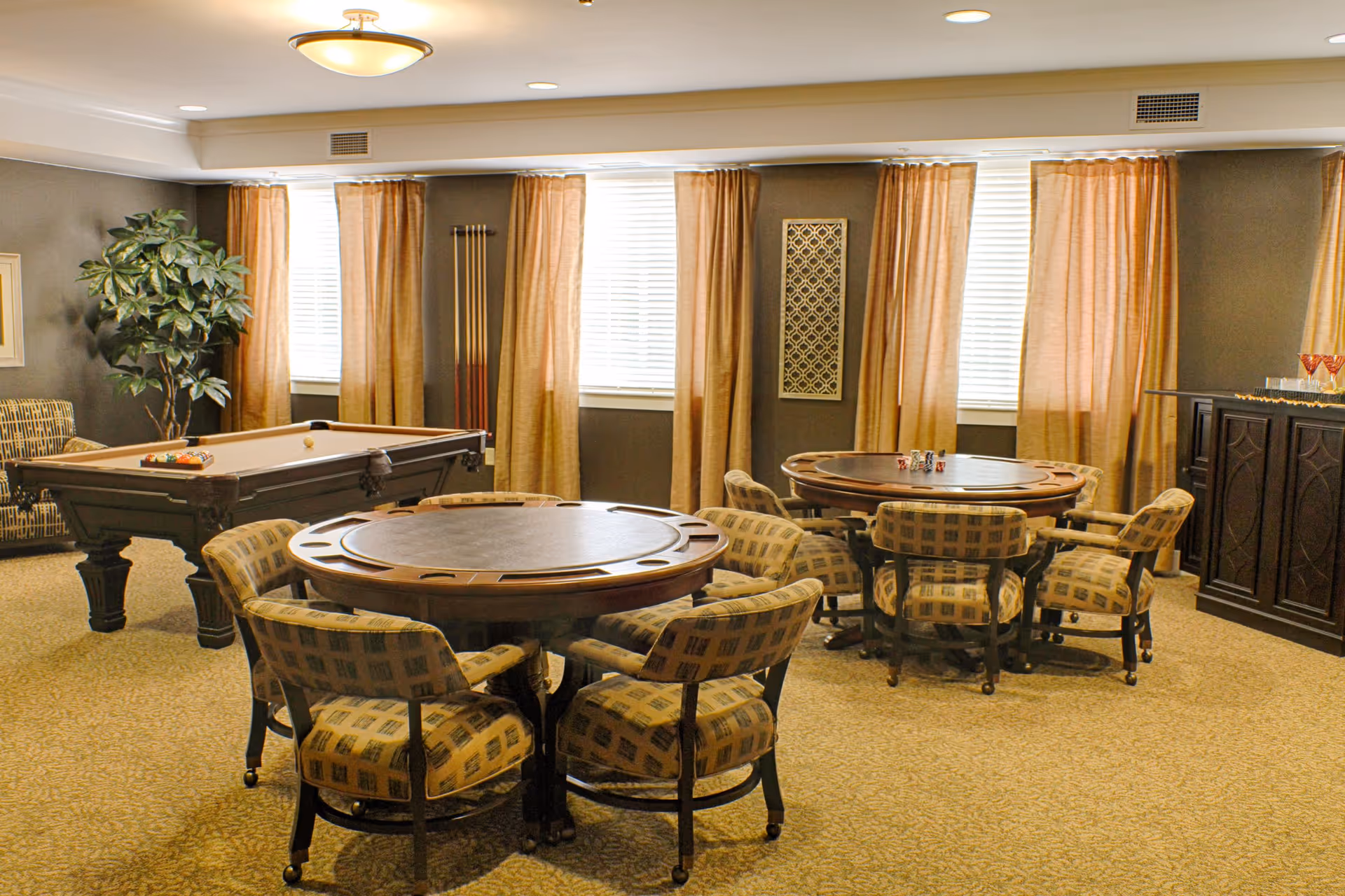 A cozy game room with two round tables surrounded by cushioned chairs, a pool table with billiard balls and cues, a potted plant, and a cabinet with glasses on top. The room has beige carpet, brown walls, and windows with light brown curtains.