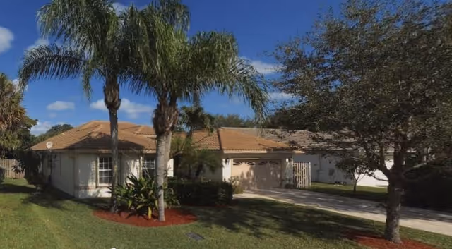Single-story residential building with a tan tiled roof surrounded by palm trees and other greenery under a blue sky with some clouds.