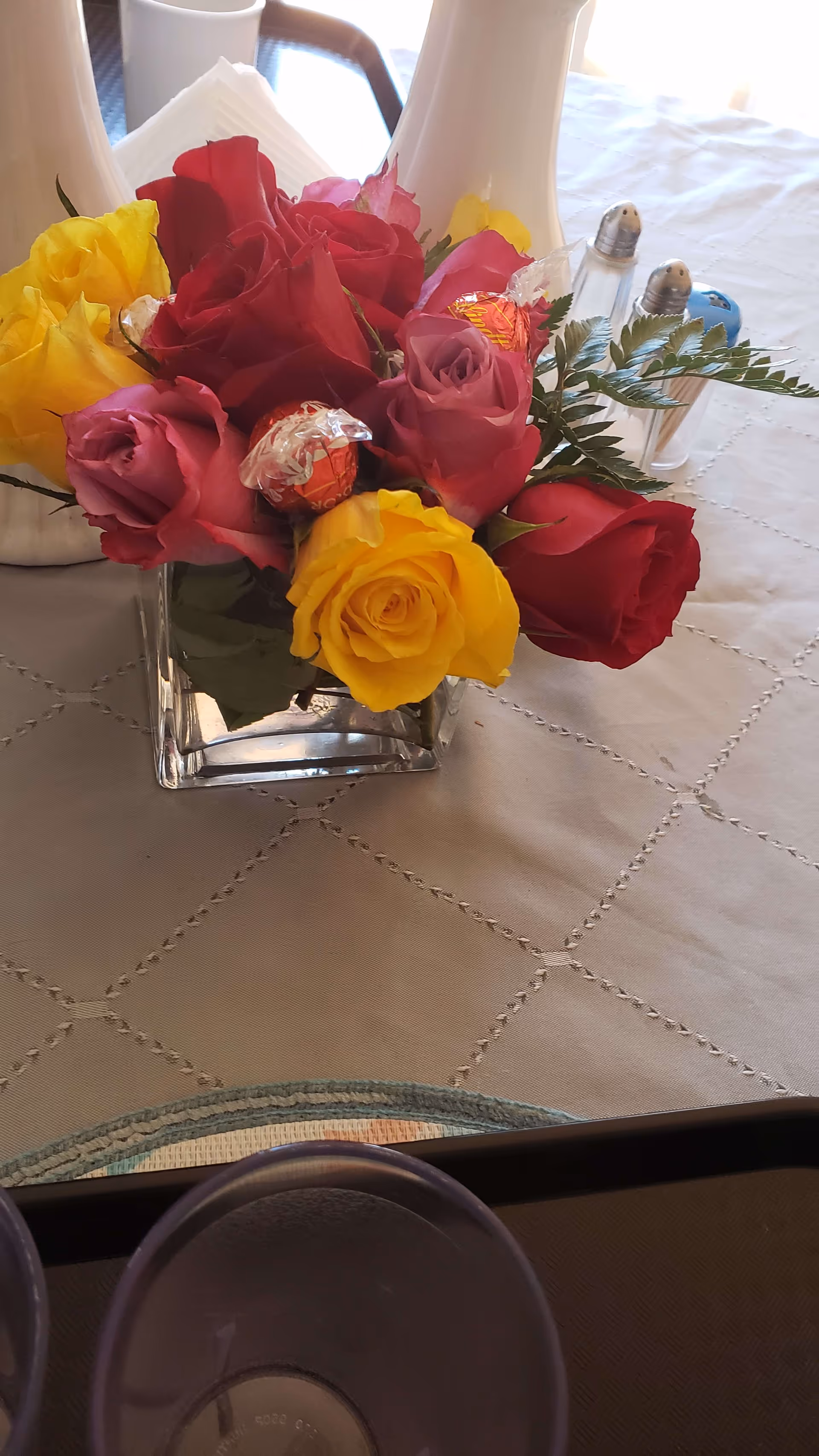 A glass vase of red, yellow, and pink roses arranged on a tablecloth next to salt and pepper shakers.