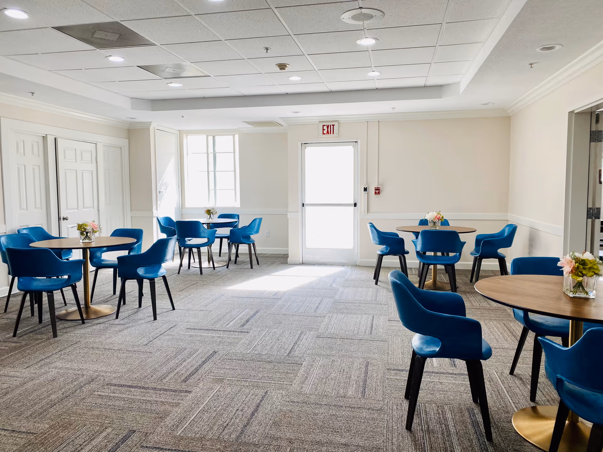 A bright and clean dining area with several round wooden tables surrounded by blue upholstered chairs. Each table has a small floral arrangement. The room has beige walls, a patterned carpet, a white ceiling with recessed lighting, and a door with an exit sign above it at the far end.