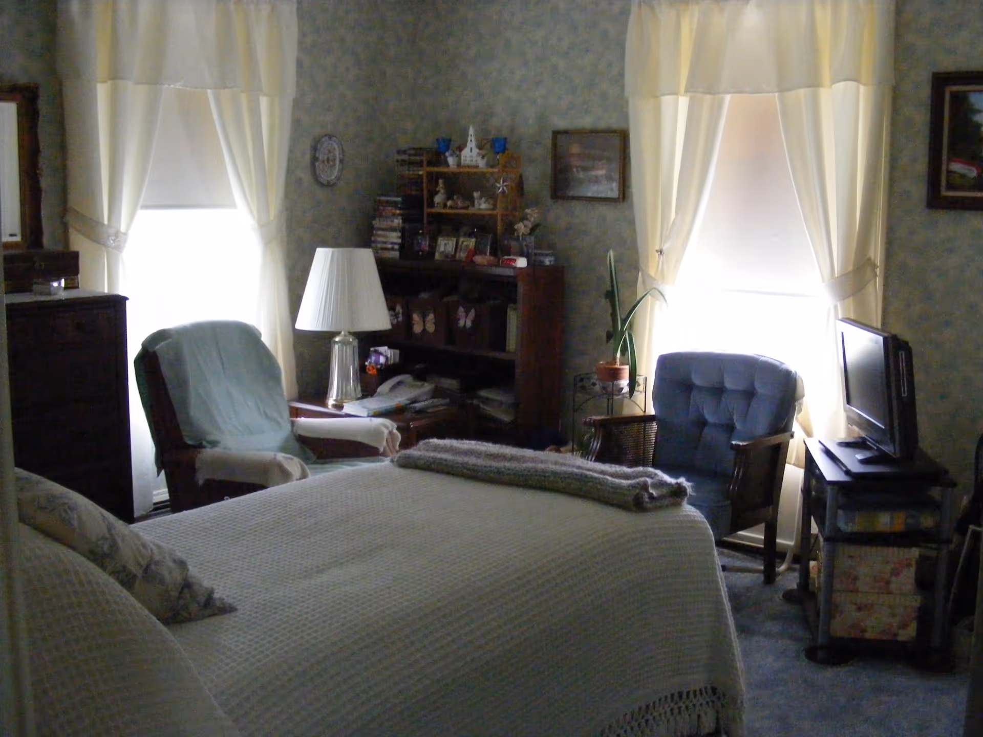 A cozy bedroom with a bed covered in a white textured blanket and pillows, two armchairs near two windows with white curtains, a small table with a lamp, a bookshelf filled with various items, and a TV on a stand.