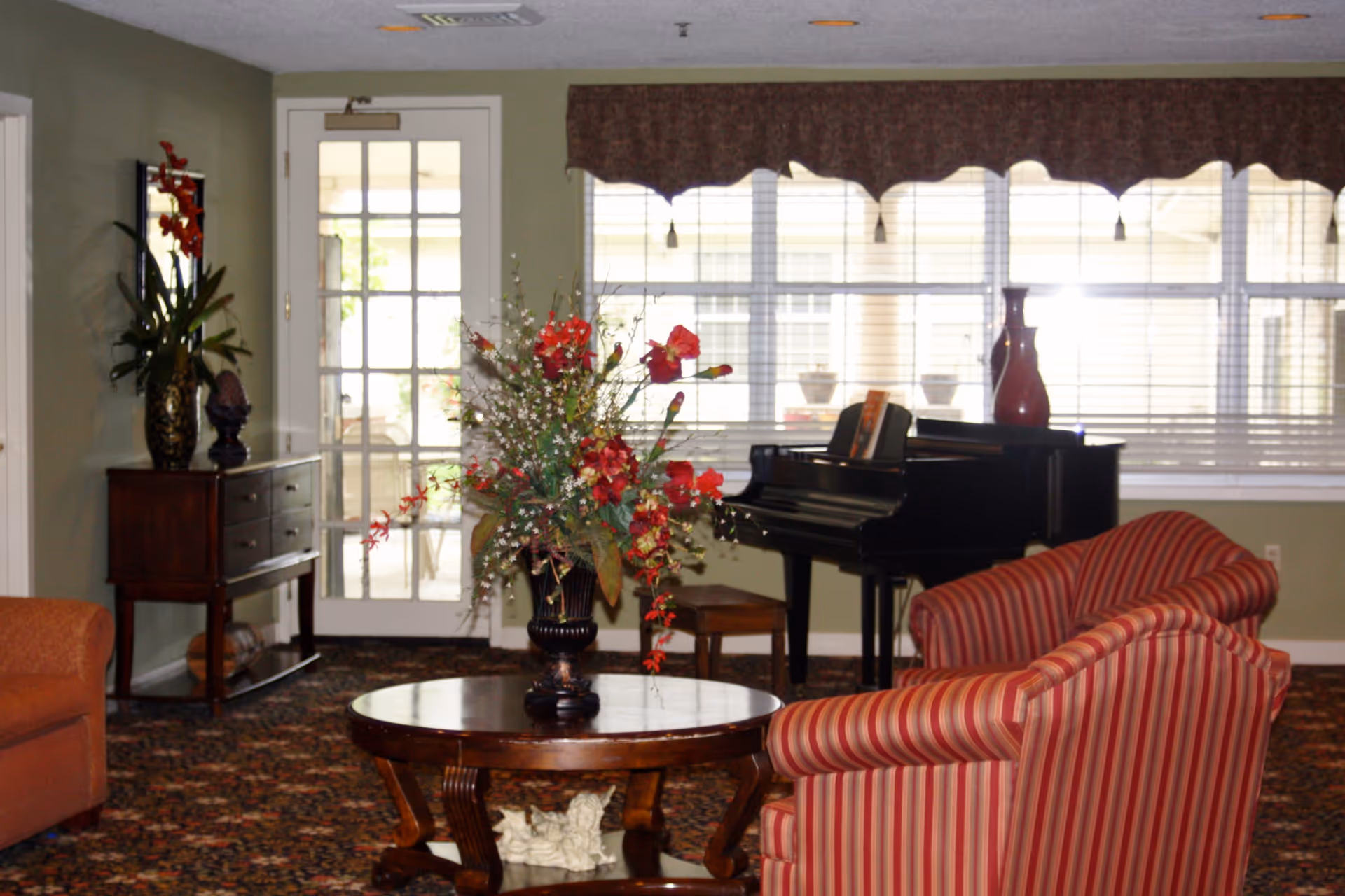 Sunlit common room with striped armchairs, a round coffee table holding a floral arrangement, and a black piano by large windows.