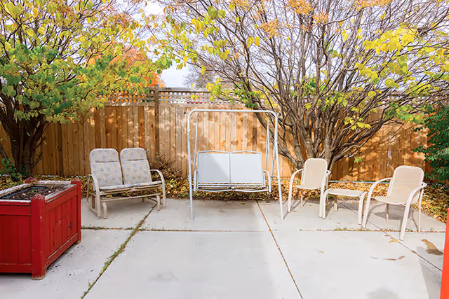 Outdoor patio area with a concrete floor, featuring a red planter box on the left, a cushioned two-seater bench, a white metal swing bench, and two beige chairs arranged near a wooden fence with trees showing autumn foliage in the background.