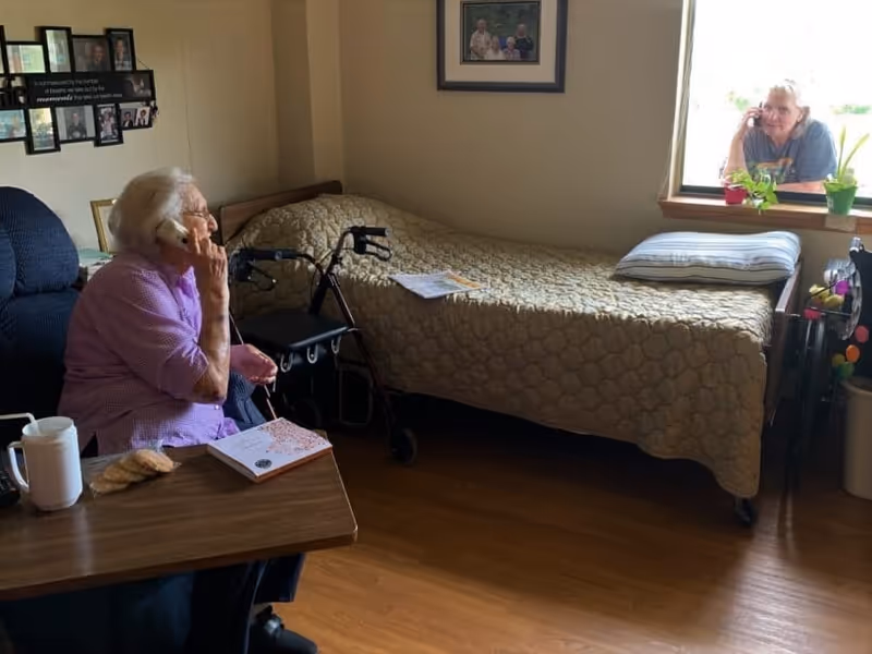 An elderly woman sitting in a chair inside a room, talking on a phone. There is a bed with a beige quilt and a pillow next to her, a walker beside the bed, and a woman outside the window also talking on a phone. The room has wooden flooring and framed pictures on the walls.