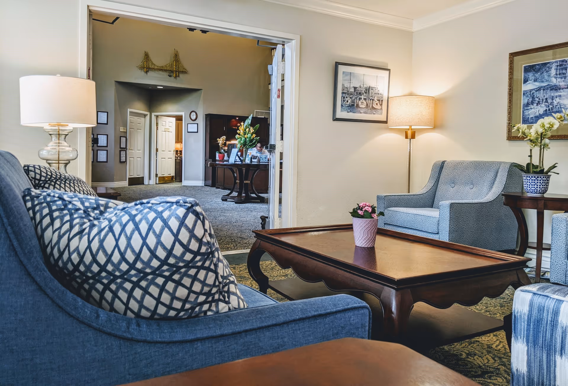 A cozy living room area with blue upholstered chairs, a wooden coffee table with a small potted plant, and two lamps providing warm lighting. The room opens into a hallway with doors and a table with flowers and decorations.