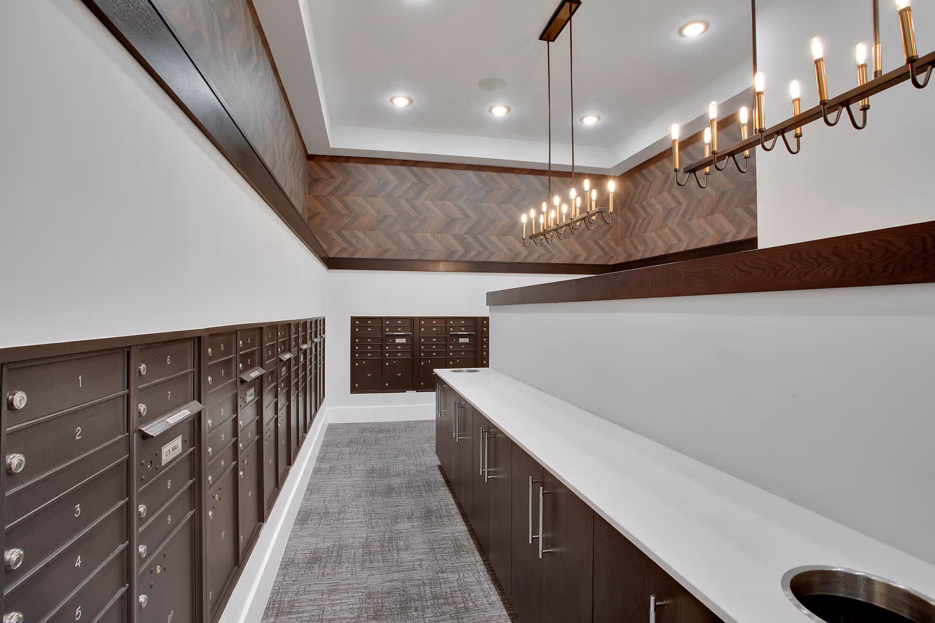 Interior view of a mailroom with multiple dark brown mailboxes mounted on the walls and a long white countertop with dark brown cabinets underneath. The room features a patterned wood accent wall near the ceiling and modern hanging light fixtures.