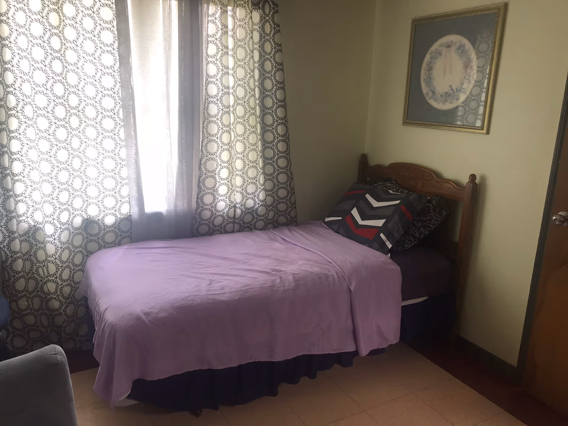 A small bedroom with a single bed covered with a purple blanket and multiple pillows. The bed is positioned next to a window with patterned curtains. There is a framed picture hanging on the wall above the bed and a closed wooden door to the right.