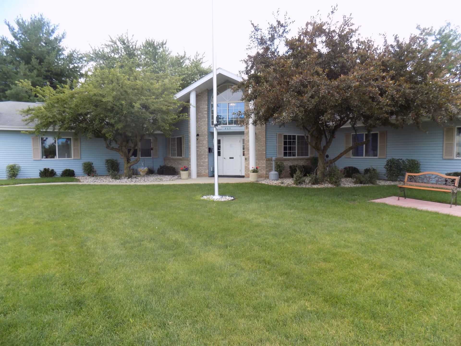 Front exterior view of a single-story building with light blue siding and beige shutters, surrounded by green grass and trees. There is a flagpole in the center of the lawn and a bench on the right side near a paved area.