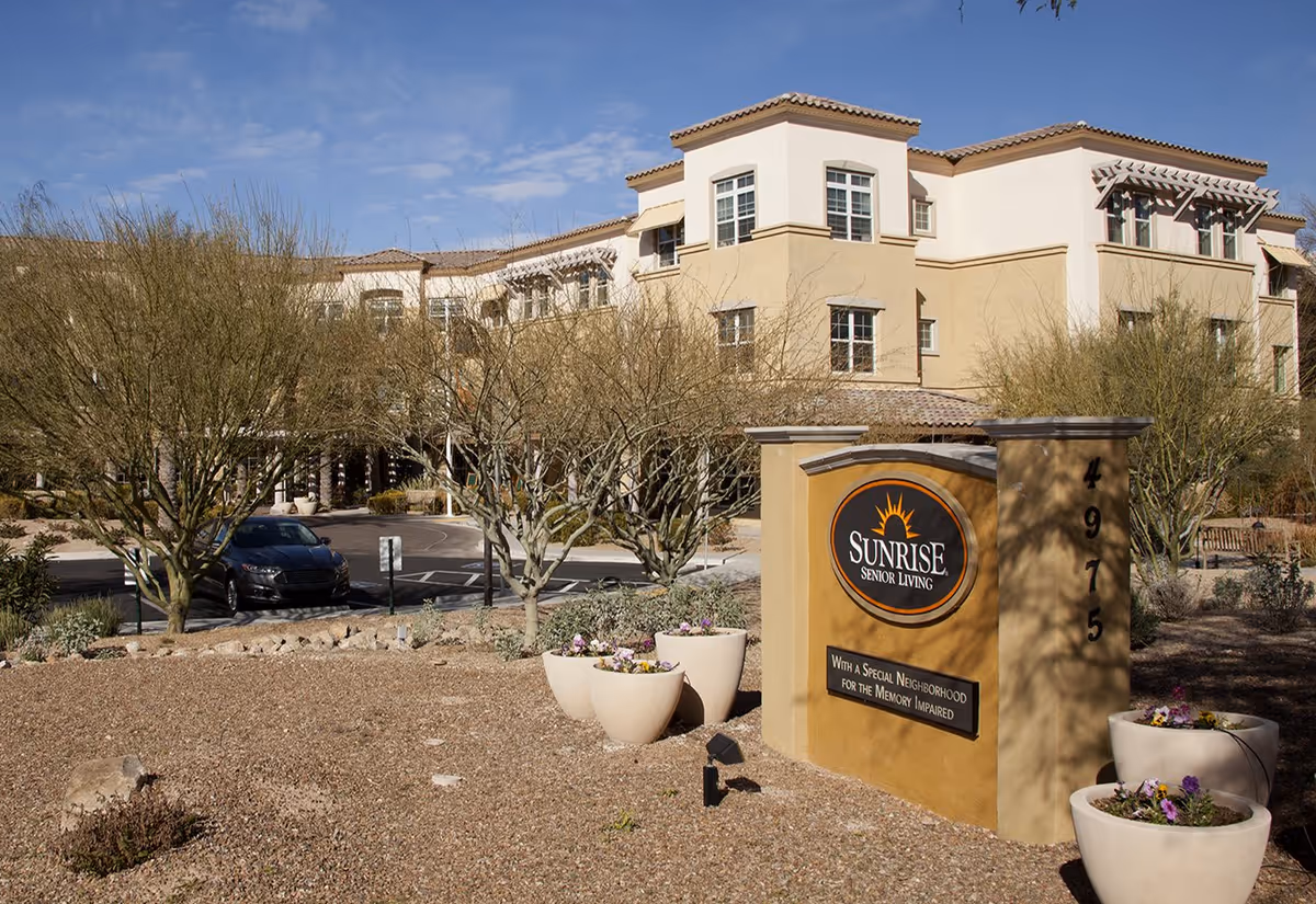 Exterior view of Sunrise Senior Living facility with a sign in the foreground that reads 'Sunrise Senior Living, With a Special Neighborhood for the Memory Impaired.' The building is beige with multiple windows and surrounded by desert landscaping including trees and flower pots.
