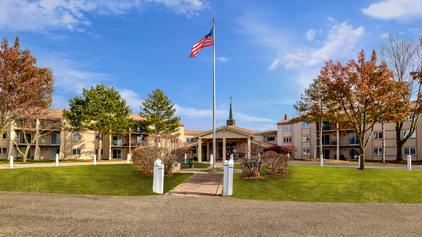 Exterior view of a senior living facility with a central entrance, a flagpole with an American flag, green lawns, trees with autumn foliage, and a clear blue sky.