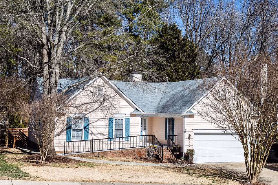 Beige home with blue shutters, brick steps, ramp railing, and a white two car garage.