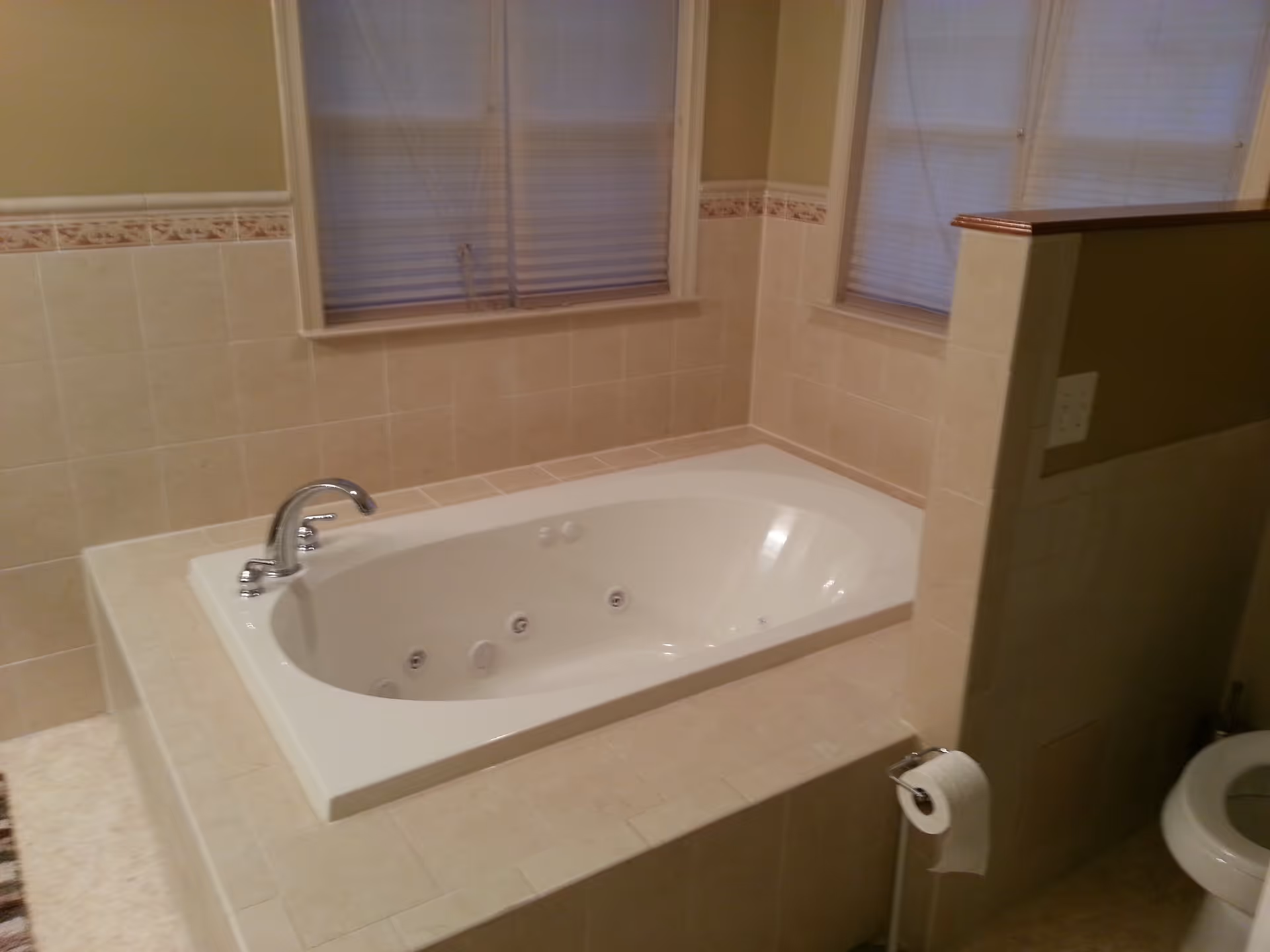 Tiled bathroom featuring a built-in jetted bathtub beneath two windows with a partially visible toilet and toilet paper holder.
