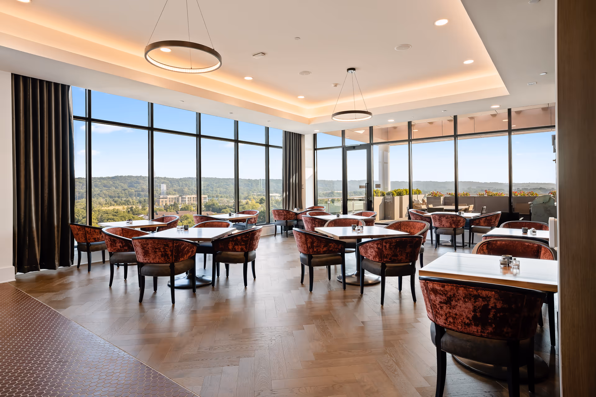 A bright dining room with large floor-to-ceiling windows offering a scenic view of greenery and hills outside. The room features multiple square tables with plush red velvet chairs arranged neatly. Modern circular pendant lights hang from the ceiling, and there is a balcony area visible through glass doors on the right side.