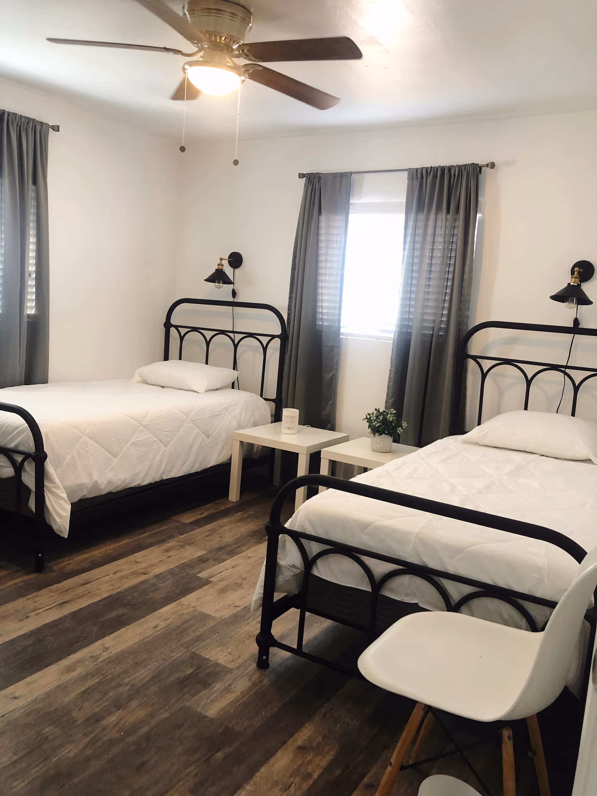 A bright bedroom with two single black metal frame beds, each made with white bedding and a pillow. Between the beds are two small white tables, one with a small plant and the other with a decorative item. The room has wood-patterned flooring, two windows with gray curtains, and a ceiling fan with a light. A white chair with wooden legs is positioned near one of the beds.
