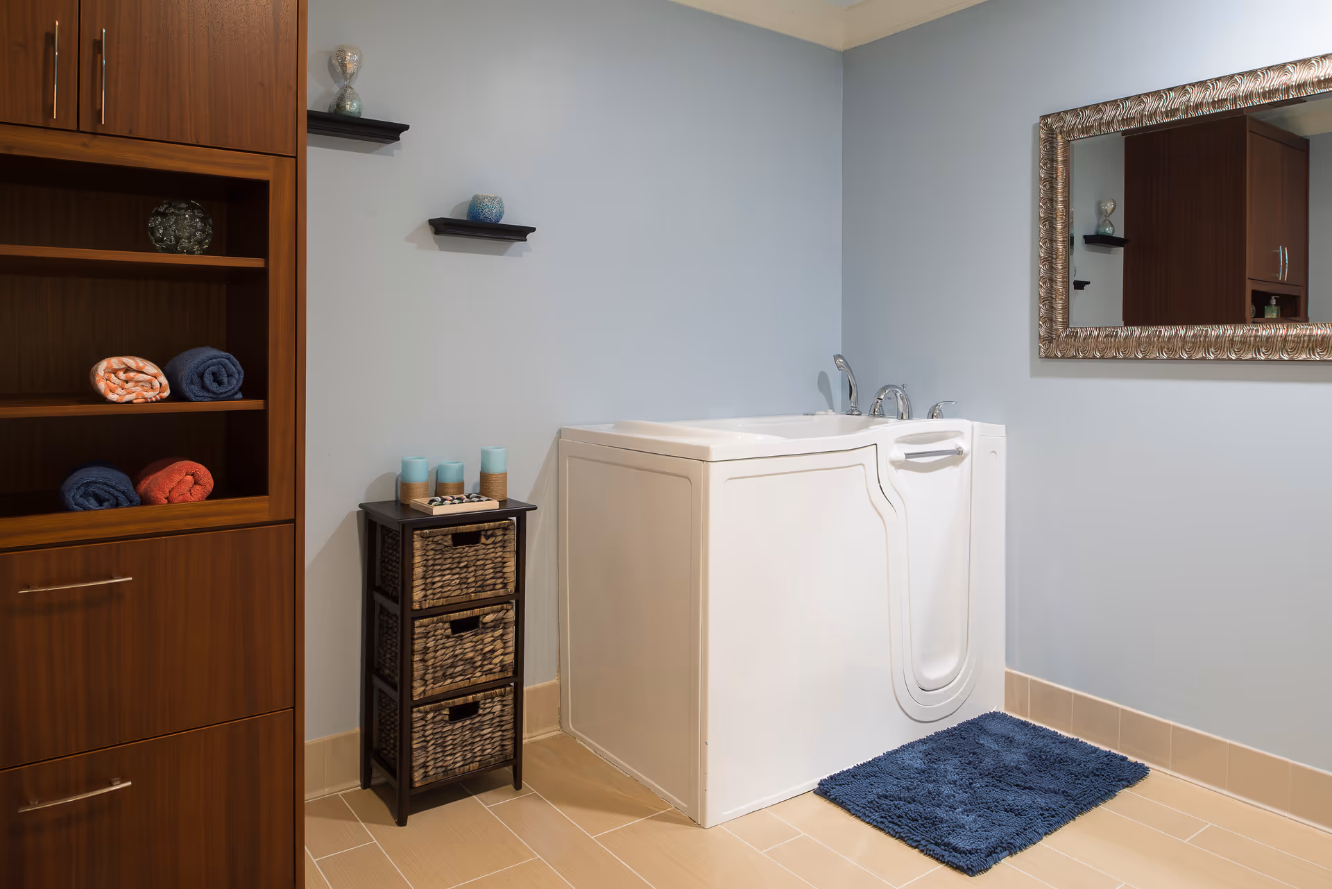 A bathroom with a walk-in bathtub featuring a door and faucet, a blue bath mat on the floor, a wooden shelving unit with rolled towels, a small cabinet with three wicker drawers topped with candles, and a large ornate mirror on the wall.
