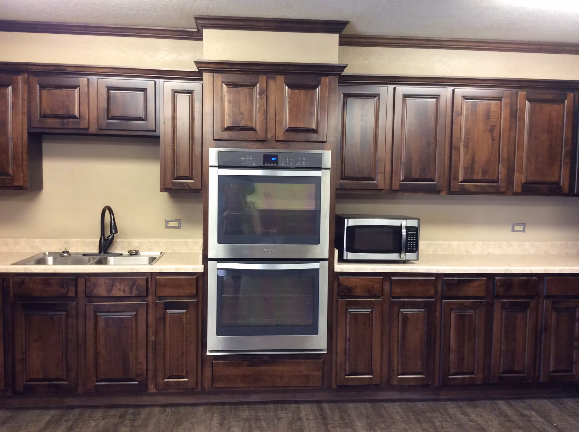 A kitchen with dark wood cabinets, a double stainless steel wall oven, microwave, sink, and beige countertops.