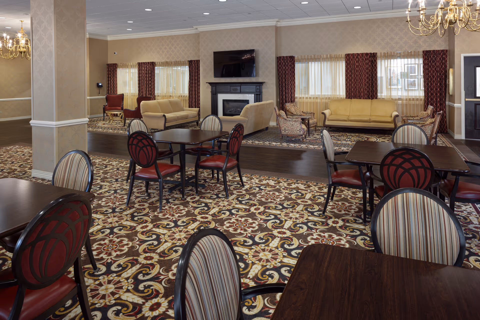 A spacious and elegant common area in a senior living facility featuring multiple dark wood tables with chairs that have red and striped upholstery. The floor is covered with a patterned carpet in shades of brown, beige, and cream. In the background, there are beige sofas and patterned armchairs arranged around a black fireplace with a mounted flat-screen TV above it. The windows are adorned with sheer curtains and dark red drapes, and chandeliers hang from the ceiling providing warm lighting.