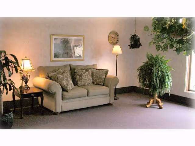 A cozy living room area with a beige loveseat adorned with patterned cushions, a wooden side table with a decorative lamp, a floor lamp, a wall clock, framed artwork, and several green plants near a window.