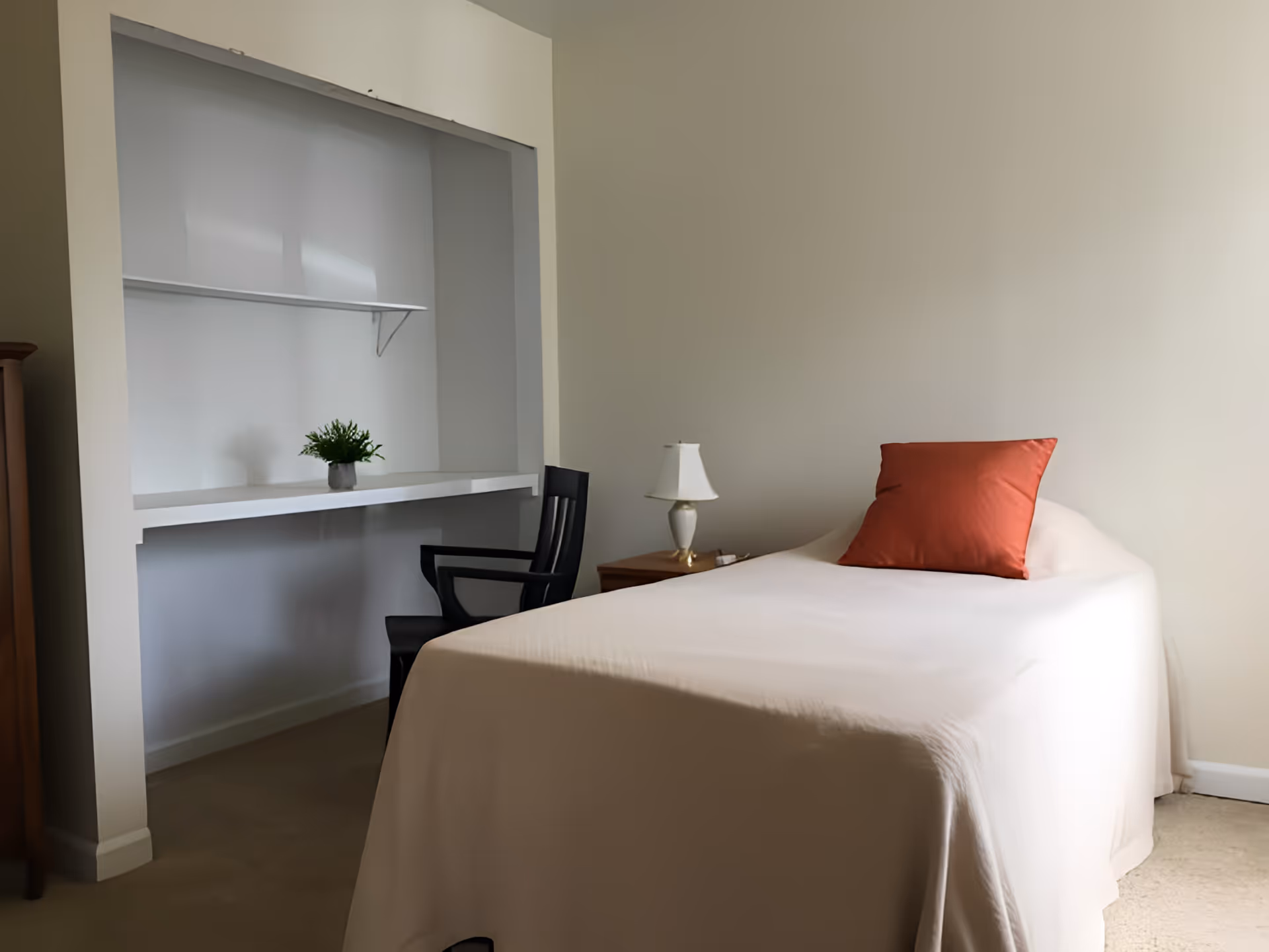 A simple bedroom with a single bed covered in a beige bedspread and a red pillow. Next to the bed is a wooden nightstand with a white lamp. There is a black chair positioned under a built-in white desk with a small green plant on it. The walls are painted light beige.