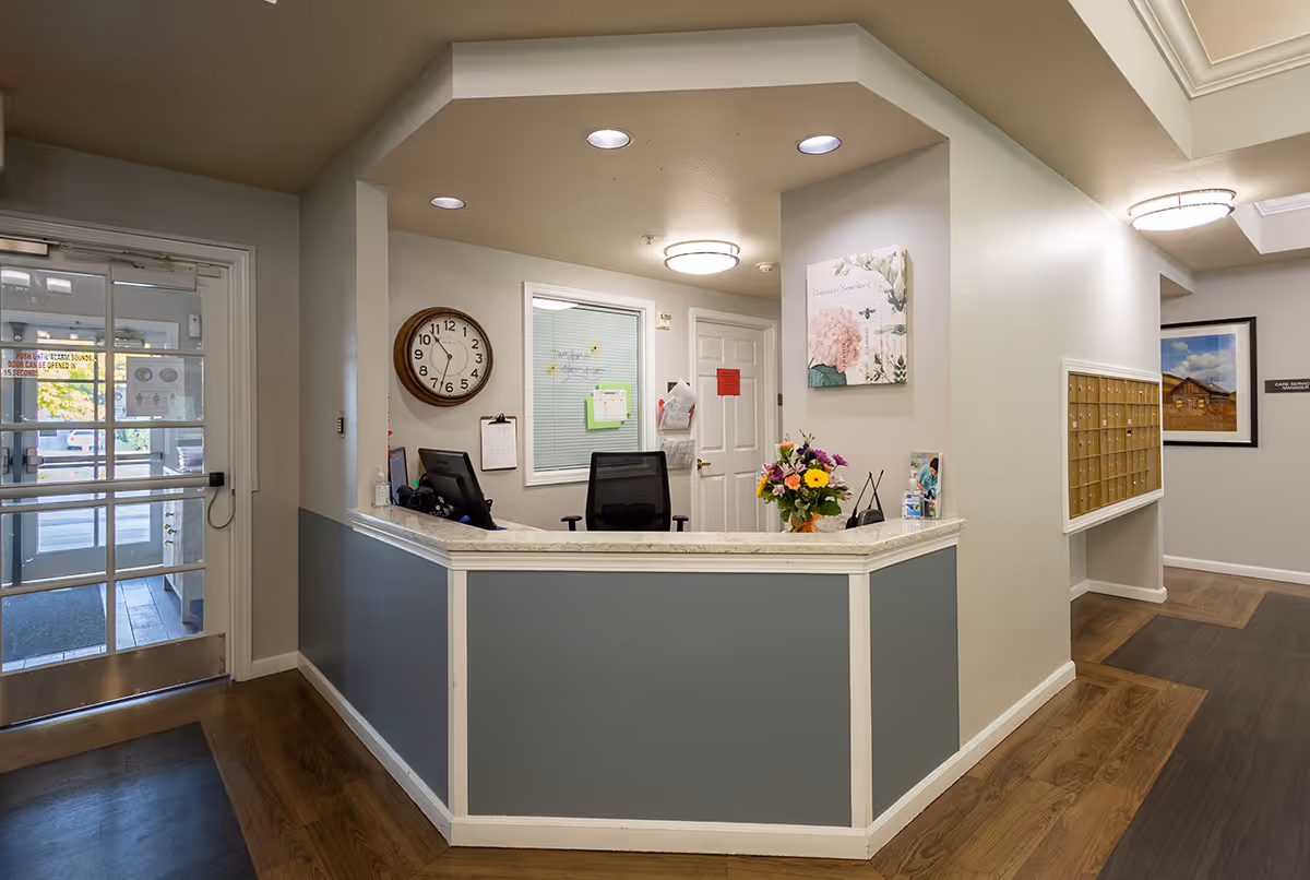 Reception area inside Avista Senior Living Yakima with a front desk featuring a computer, office chair, and a vase of flowers. A large clock is mounted on the wall behind the desk, and there is a door and a window with blinds. The entrance door with glass panels is visible to the left, and a set of mailboxes is mounted on the wall to the right.