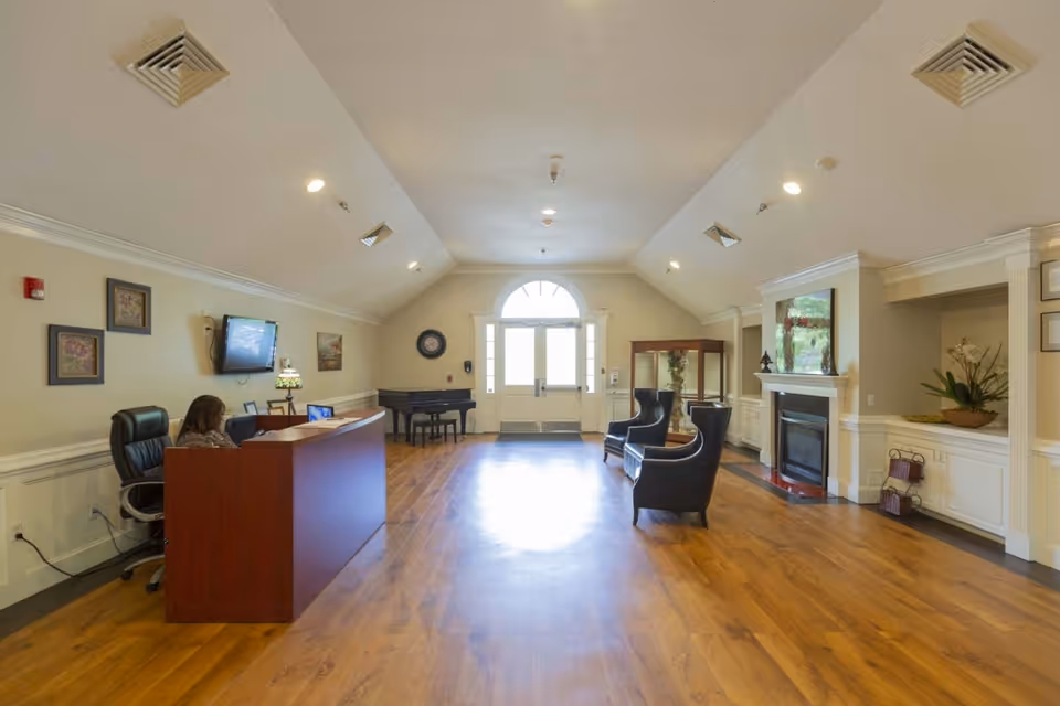 A spacious, well-lit reception area with wooden flooring and a high ceiling. On the left, a woman is seated behind a wooden reception desk with a computer. The room features a wall-mounted TV, framed artwork, a piano in the back corner, two black armchairs near a fireplace on the right, and a large windowed door letting in natural light at the far end.