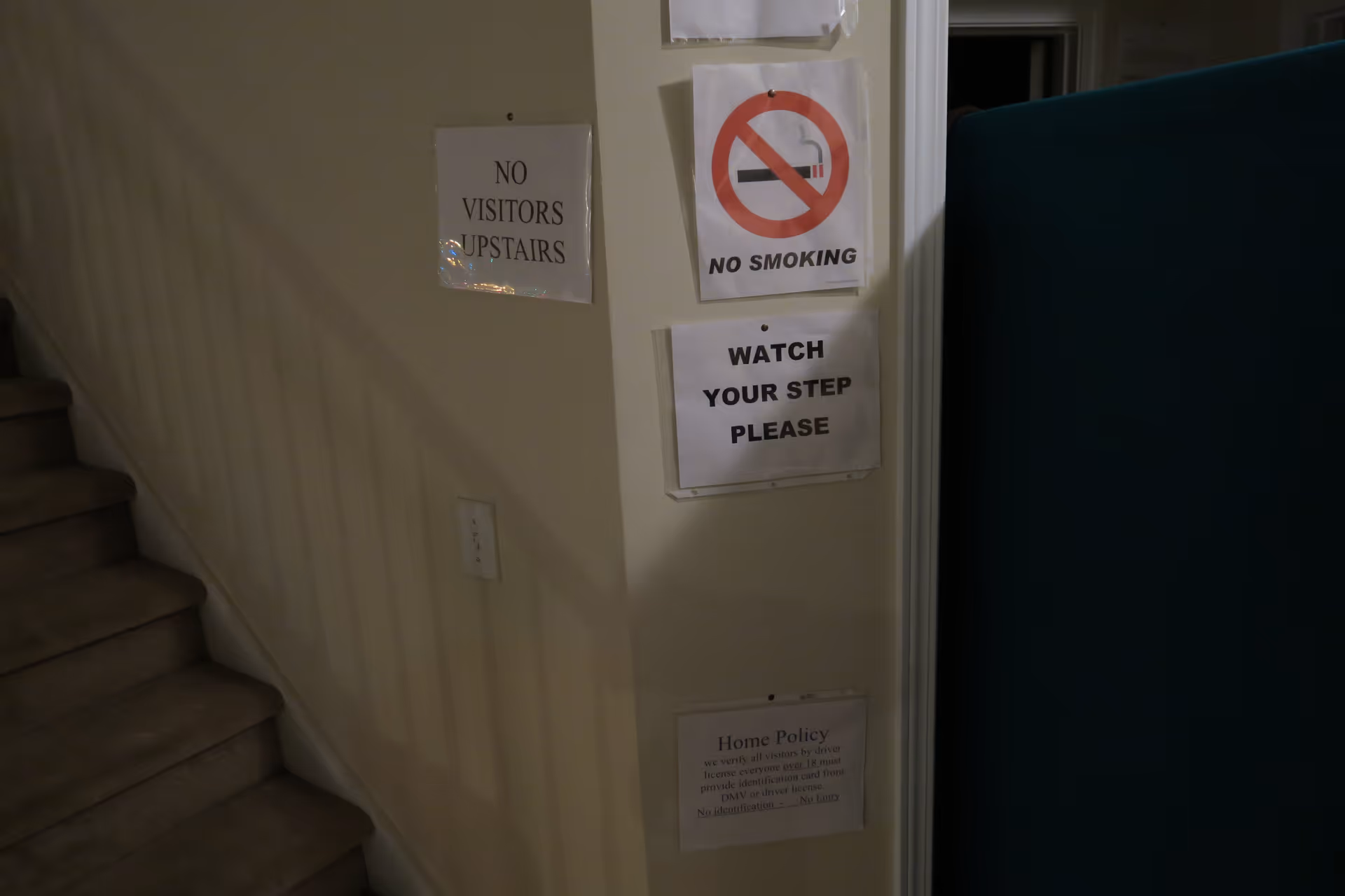A dimly lit indoor corner showing a carpeted staircase on the left and a wall with four posted signs. The signs read: 'NO VISITORS UPSTAIRS', a no smoking symbol with 'NO SMOKING', 'WATCH YOUR STEP PLEASE', and a home policy notice about visitor identification requirements. A blue partition or piece of furniture is partially visible on the right.