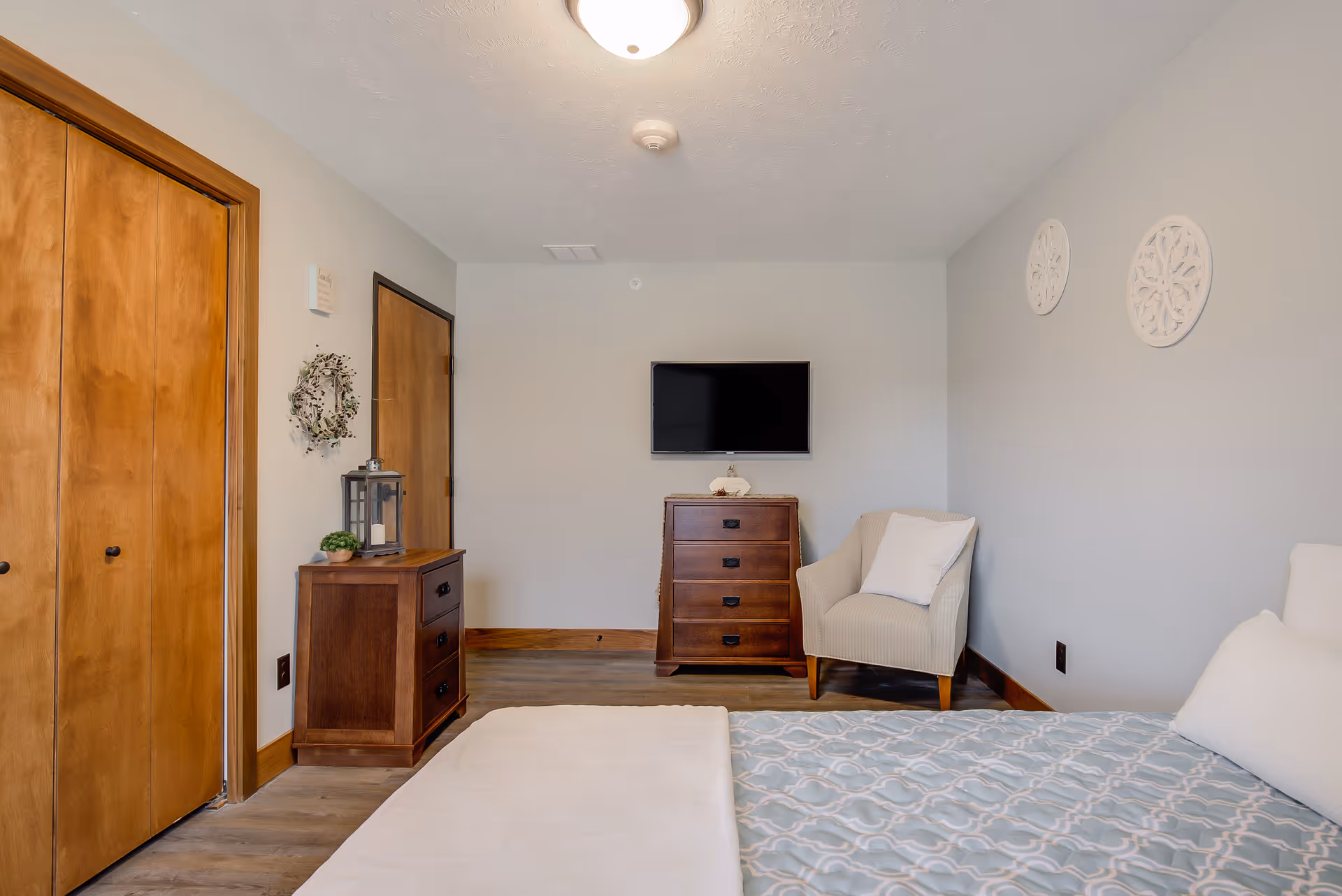A bedroom with a bed in the foreground covered with a patterned blue and white blanket. There is a wooden dresser with a flat-screen TV mounted on the wall above it. To the right of the dresser is a beige armchair with a white pillow. On the left side of the room, there are two wooden doors and a small wooden nightstand with decorative items including a lantern and a small plant. The walls are painted light gray with two round white wall decorations on the right wall.