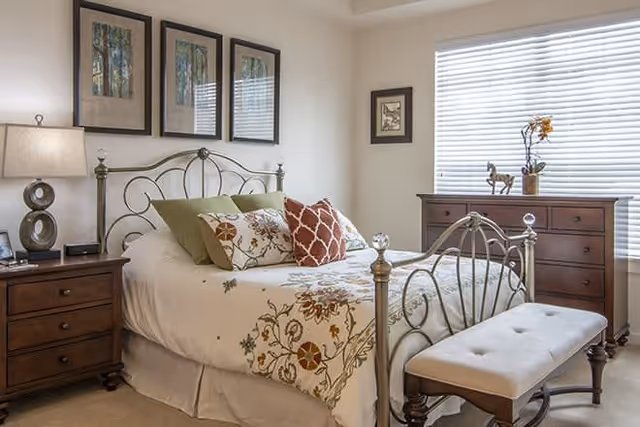 A cozy bedroom with a metal frame bed adorned with floral and patterned pillows and a matching bedspread. There is a wooden nightstand with a lamp and a clock on the left side of the bed. On the right side, there is a wooden dresser with decorative items including a small sculpture and a vase with flowers. A cushioned bench sits at the foot of the bed. The room has three framed pictures above the bed and a window with blinds letting in natural light.