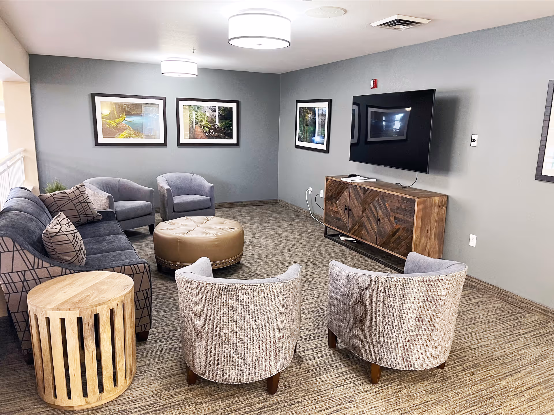 A cozy senior living facility lounge area with a gray sofa, two gray armchairs, a round beige ottoman, and a wooden side table. The walls are painted gray and decorated with framed nature photographs. A flat-screen TV is mounted on the wall above a wooden media console. The room has carpeted flooring and ceiling lights.