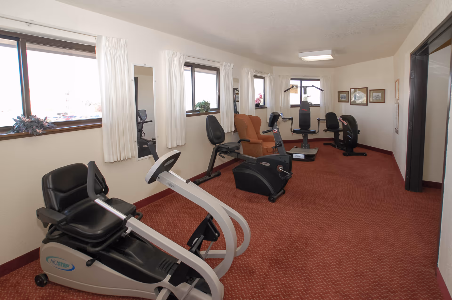 Small exercise room with recumbent bikes, a weight machine and chairs along carpeted floor under windows.