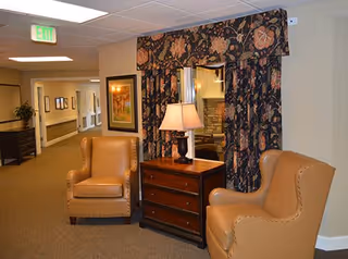 A cozy seating area in a hallway featuring two tan leather armchairs positioned around a wooden chest of drawers with a table lamp on top. Behind the chest is a window with floral-patterned curtains. The hallway extends to the left with framed artwork on the walls and an exit sign visible on the ceiling.