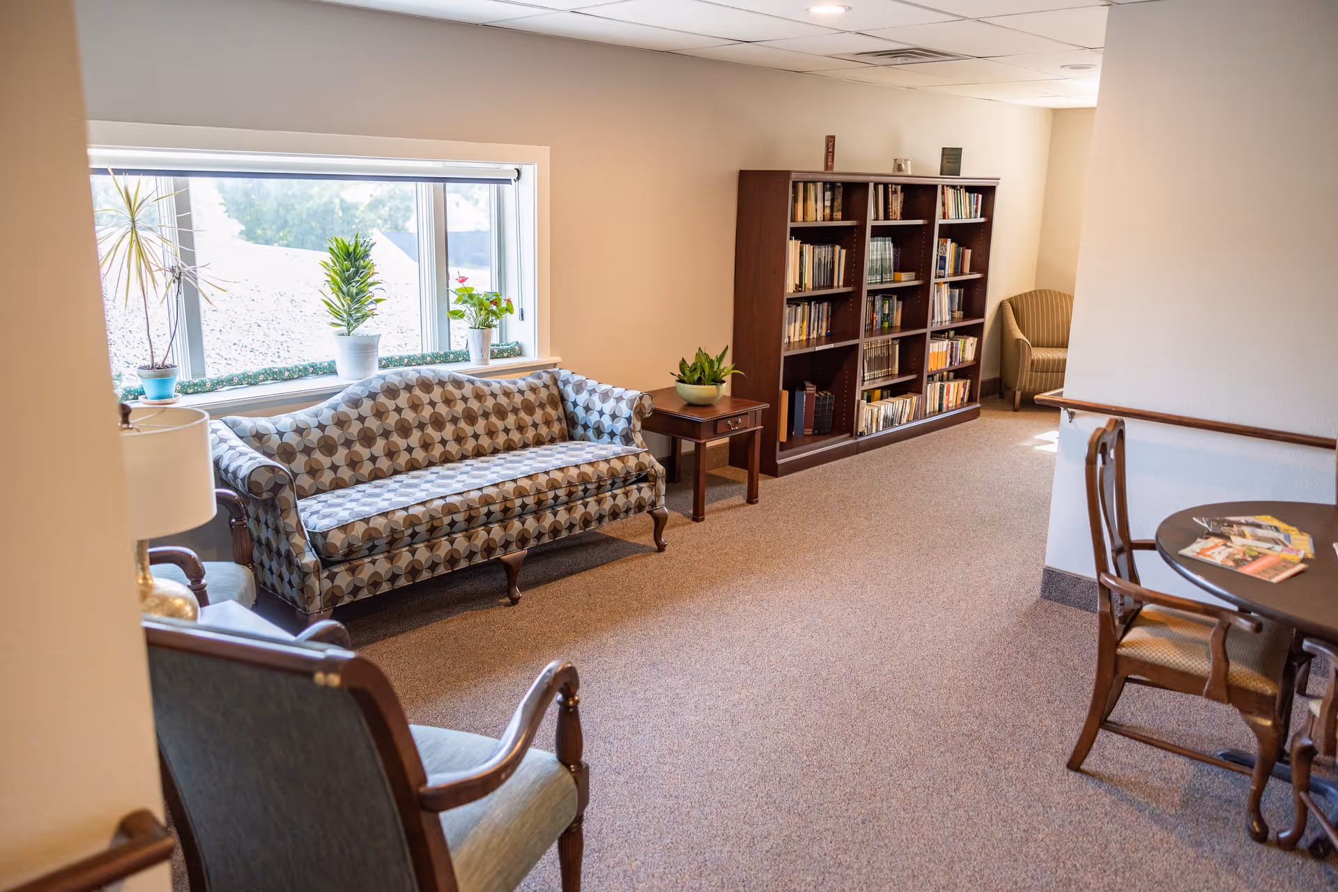 A cozy sitting area in a senior living facility with a patterned sofa, two armchairs, a wooden bookshelf filled with books, a small side table with a plant, and a round table with chairs and magazines. There is a large window with potted plants on the windowsill letting in natural light.
