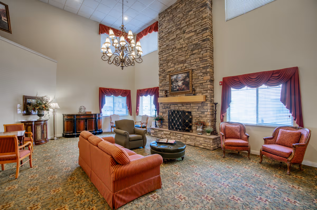 A spacious living room with high ceilings featuring a large stone fireplace with a painting above it. The room is furnished with a red striped sofa, an olive armchair, two red upholstered armchairs, and two wooden chairs with cushions. There is a round green ottoman in the center and a chandelier hanging from the ceiling. The windows have red drapes and blinds, and the carpet has a floral pattern.