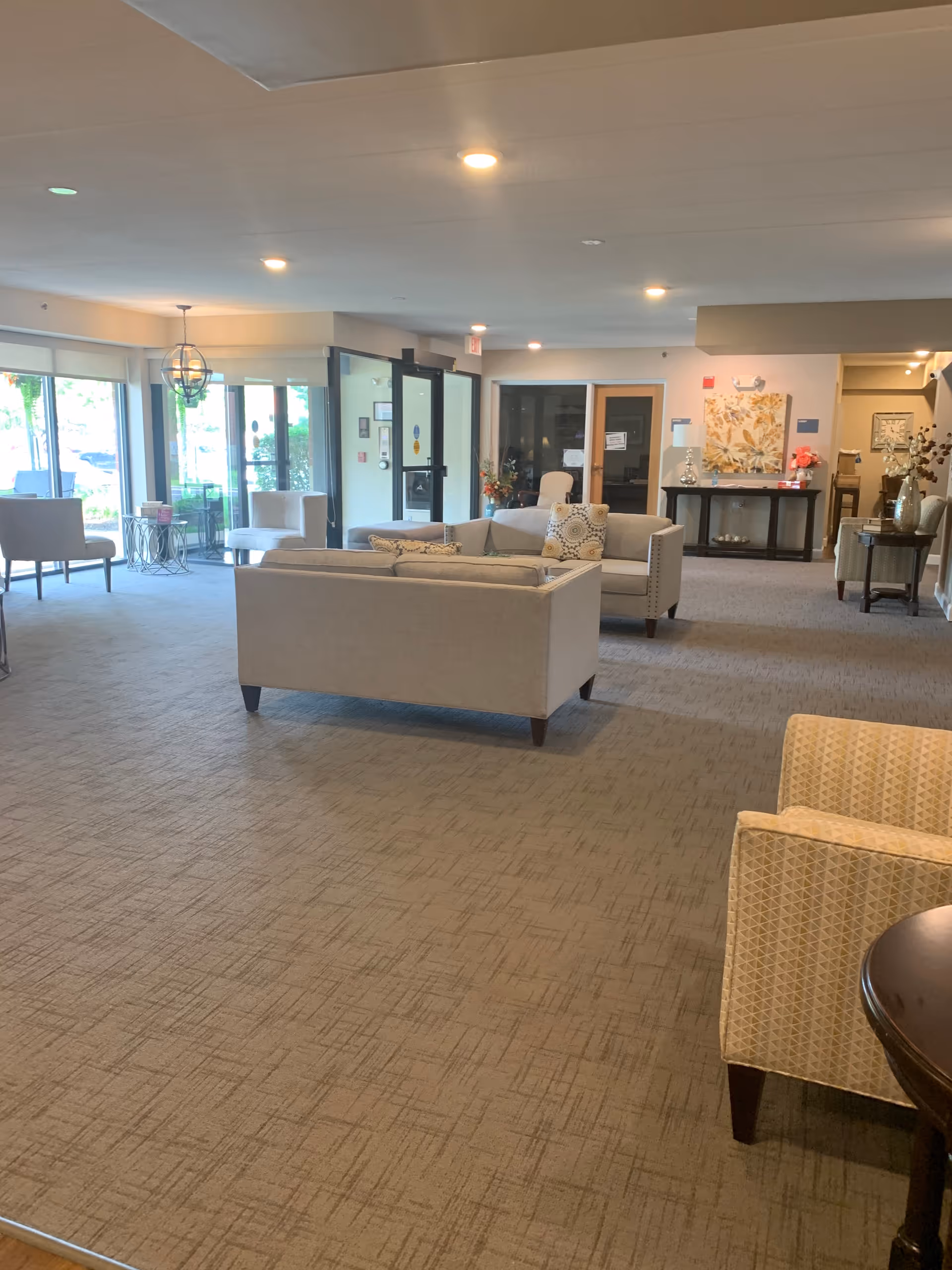 Spacious senior living lounge with sofas, armchairs, tables, and large windows near the entrance.